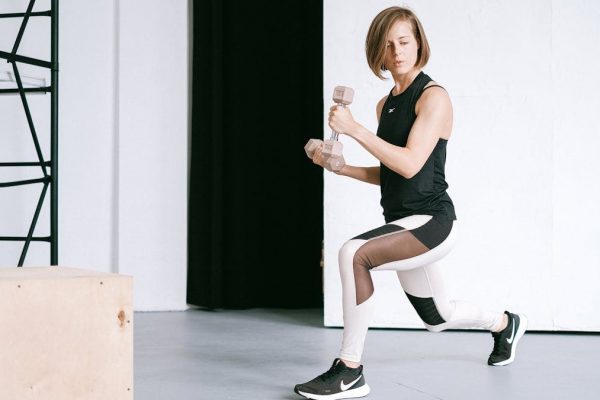 A young woman performing lunges with dumbbells in a modern indoor gym setting.